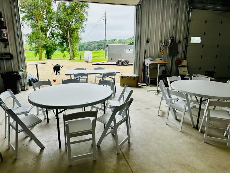 two round tables, one banquet table and chairs around each table set up in side a garage filled with garage type items