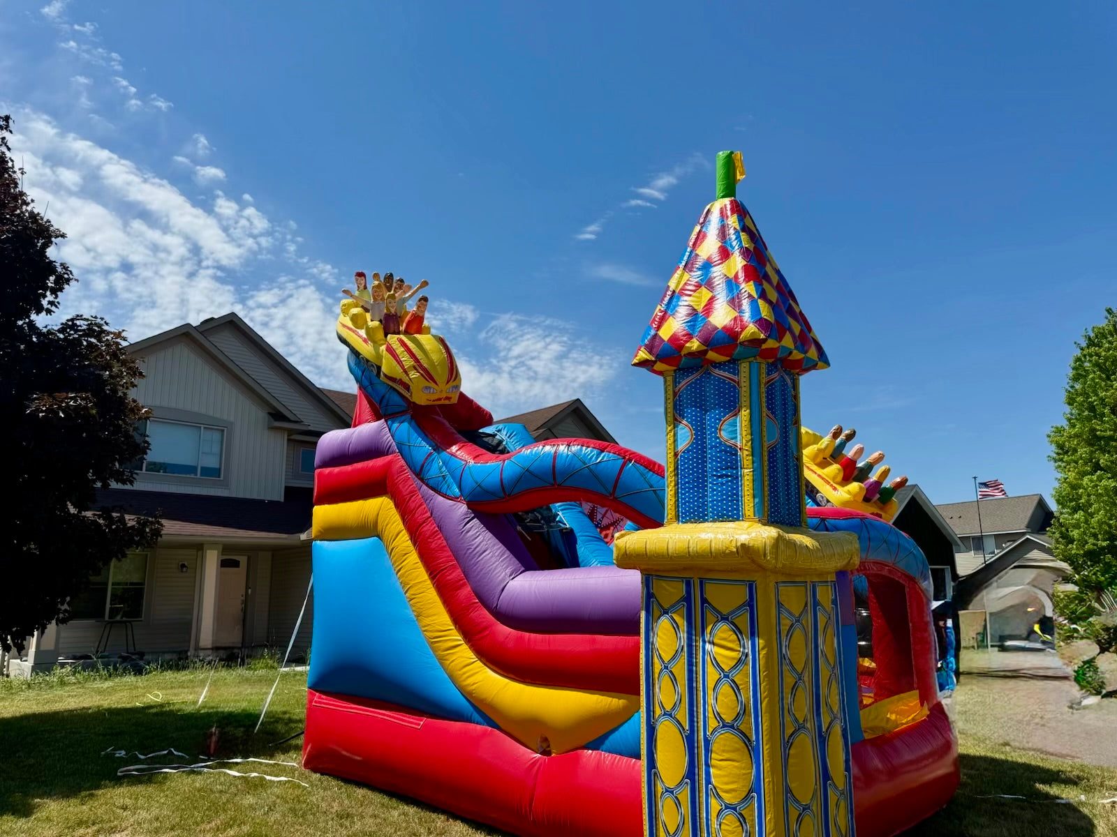 Colorful inflatable castle bounce house on a grassy area with houses and trees in the background.