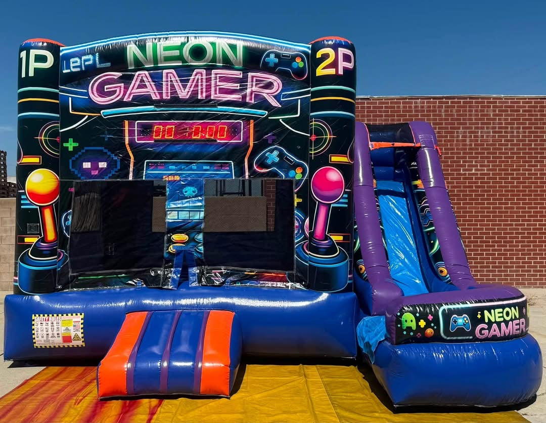A large inflatable bounce house and slide combo with a neon video game theme, featuring colorful panels, arcade-style graphics, and a purple slide, set outdoors against a brick wall and blue sky.