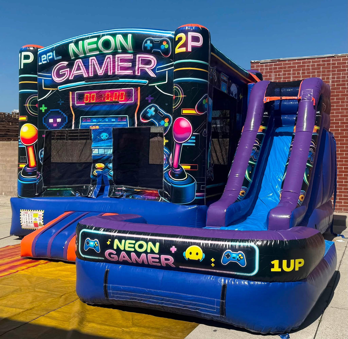 A large inflatable bounce house with a slide, decorated with neon video game graphics and the words 'NEON GAMER' and '1UP'. The structure is photographed outdoors on a paved surface with a brick building and blue sky in the background.