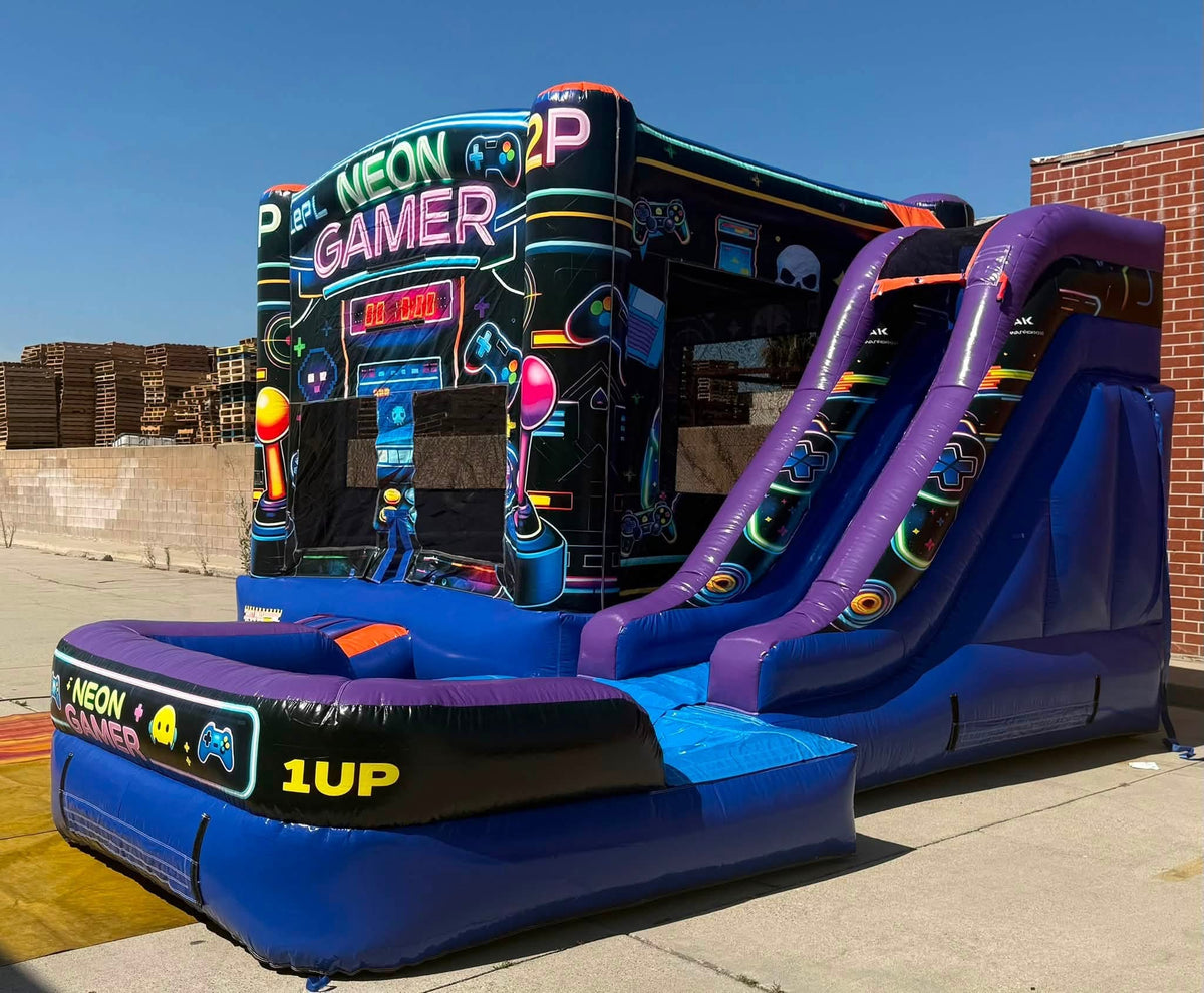A large inflatable bouncy castle with a slide and neon-style arcade graphics, photographed outdoors on a concrete surface beside a brick wall and stacked pallets under a blue sky.