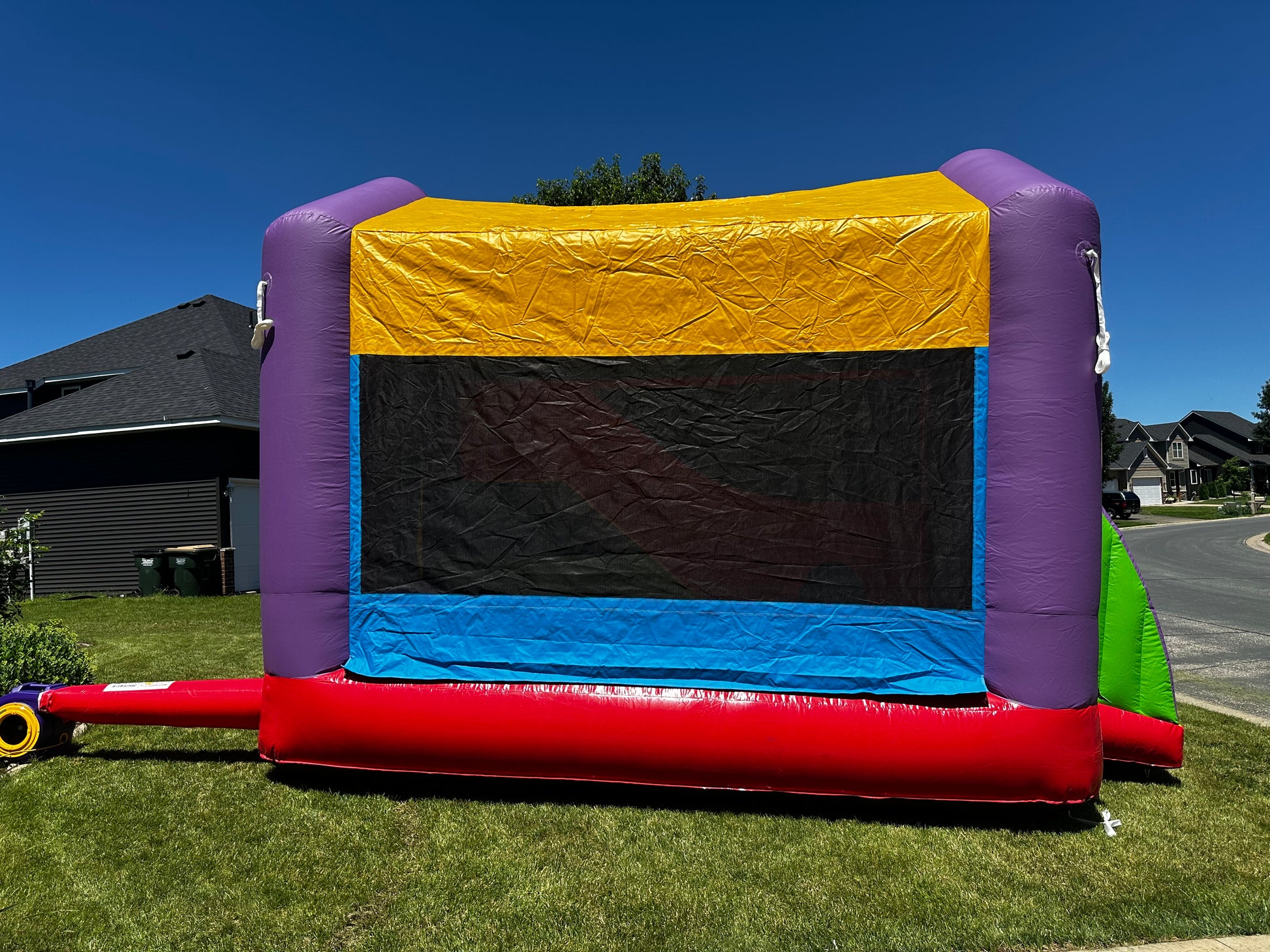 Side view of a colorful bounce house with purple, yellow, blue, and red panels, set up on a green lawn in a residential neighborhood.